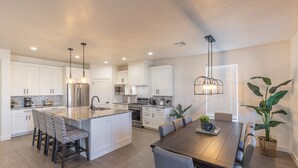 Cozinha moderna com armários brancos, uma ilha central com banquetas, mesa de jantar com cadeiras e iluminação pendente.