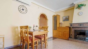 Een eetruimte met een houten tafel en stoelen, een klok aan de muur, een open haard en een klein plantje.