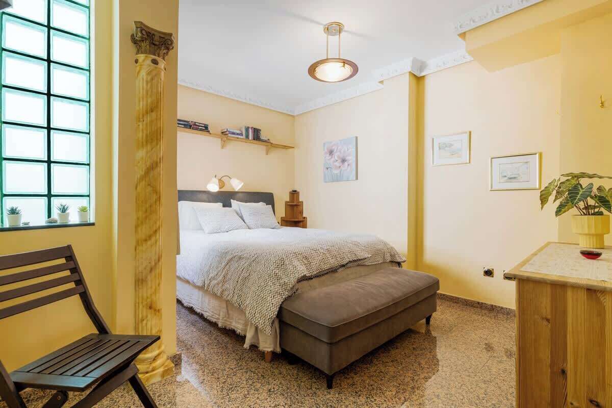 A bedroom with a bed, bench, bedside table, and a window with plants.