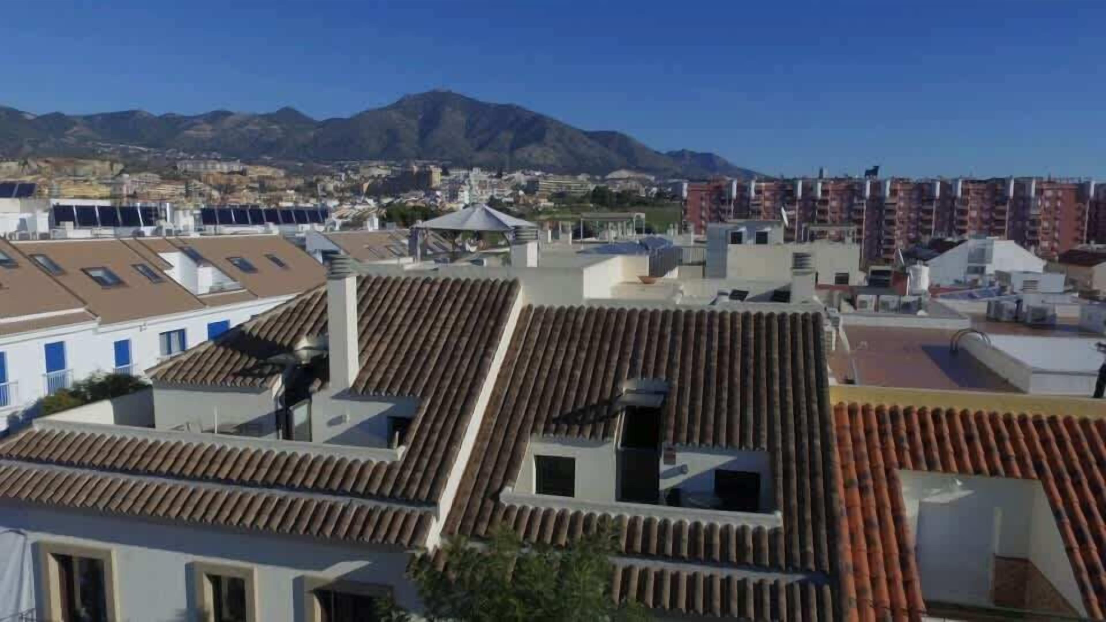 A residential area with terracotta-tiled roofs, a mix of single and multi-story buildings, and a mountainous backdrop.