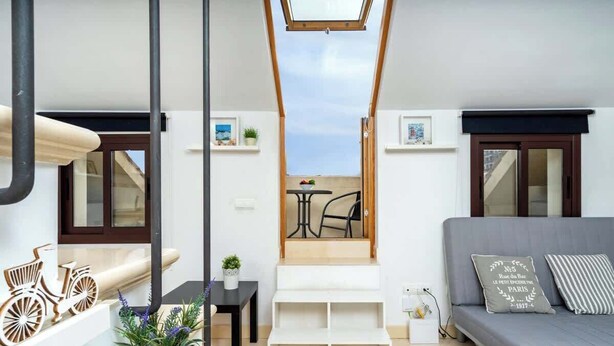 Una sala de estar moderna con un sofá gris, una pequeña mesa con una planta y vistas al exterior a través de un tragaluz.