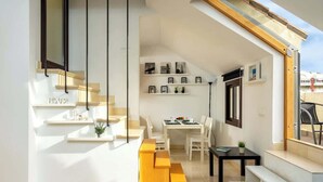 Un interior moderno con una escalera que conduce a un balcón, un comedor con mesa y sillas, y estanterías con cuadros enmarcados.