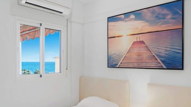 Habitación de hotel con una cama, ventana con vista al mar y un cuadro de atardecer en la pared.