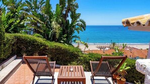 Une terrasse avec deux chaises en bois et une table, donnant sur une plage bordée de palmiers et surplombant un ciel bleu azur.