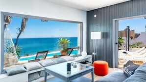 Un salon moderne doté d’une grande fenêtre donnant sur la mer, d’un canapé, d’une table basse et d’un pouf rouge.