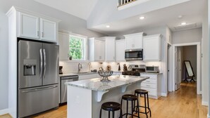 Una cocina moderna con armarios blancos, electrodomésticos de acero inoxidable, una isla central con taburetes y vistas al exterior.