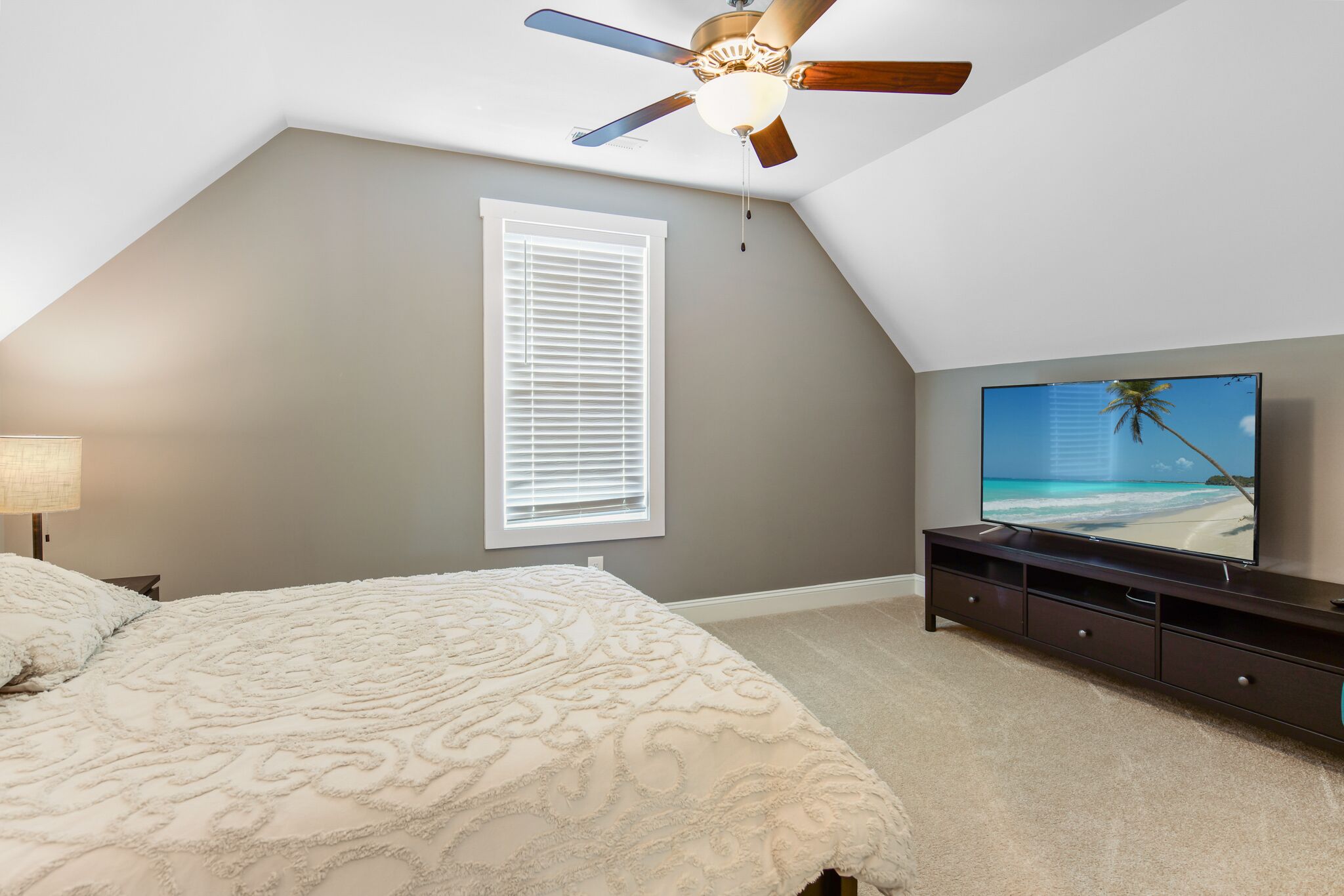 A bedroom with a ceiling fan, a large TV, a bed with a patterned bedspread, a window with blinds, and a nightstand with a lamp.