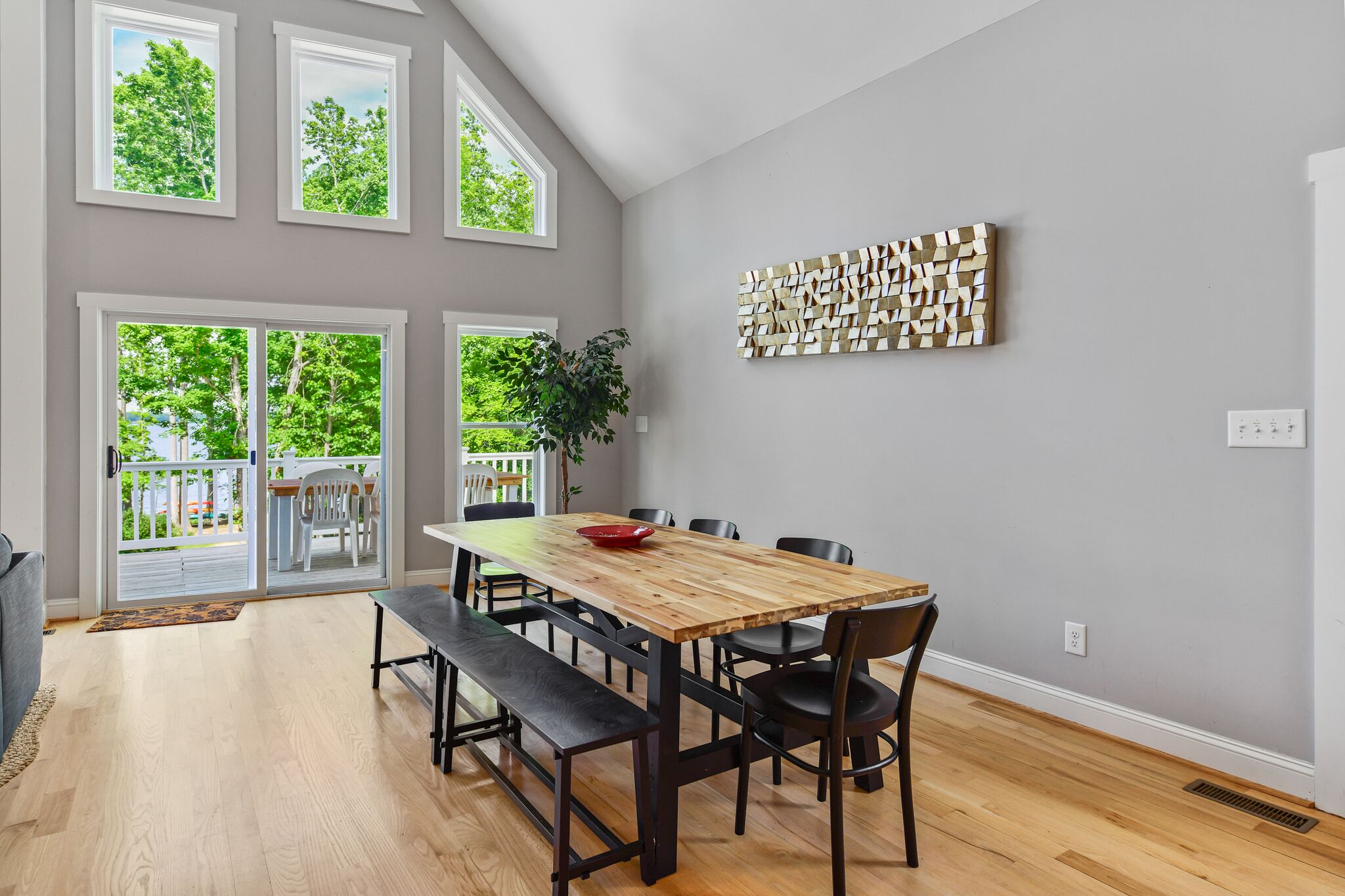 A dining area with a wooden table, black chairs, and a bench. There are large windows with a view of trees and a balcony.