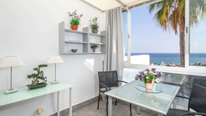 A modern room with a glass table, two chairs, a small desk with a lamp, a potted plant, and a view of the sea through a large window. - Benalmádena with sauna by Costarentals - Ref 72 (Benalmádena)