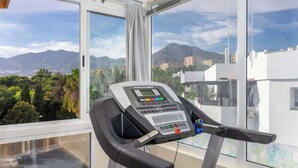 A treadmill with a digital display, located in a room with large windows offering a view of mountains and buildings. - Benalmádena with sauna by Costarentals - Ref 72 (Benalmádena)