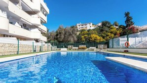 Une piscine à l’eau d’un bleu cristallin, entourée d’un bâtiment blanc doté de balcons, d’une clôture verte et offrant une vue sur des arbres et d’autres bâtiments en arrière-plan.