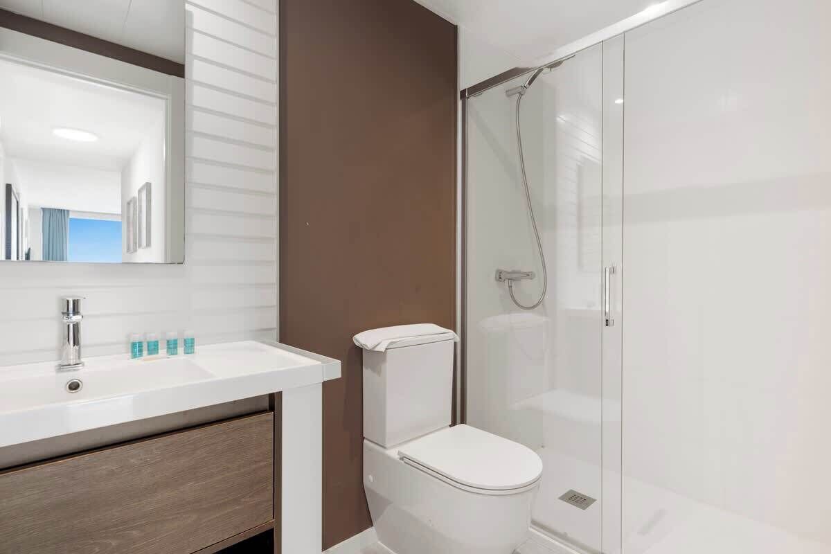 Un baño moderno con lavamanos, espejo y retrete blancos, y ducha con mampara de vidrio.