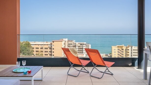A balcony with two orange chairs, a small table with a tray, and a view of the sea and buildings. - Modern Apartment w/ Pool & Gym Near Beach Access (Málaga)