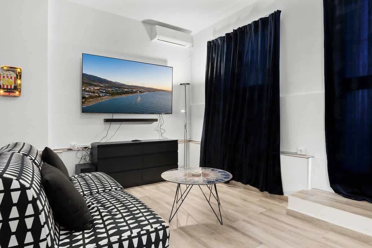 A modern living room with a large flat-screen TV, a black sofa with patterned cushions, a glass coffee table, and a view of a body of water through the curtains.