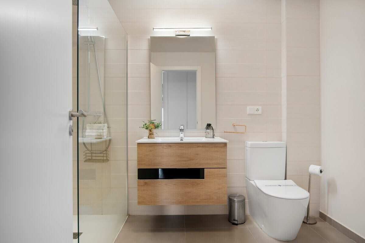 Een moderne badkamer met een glazen douchecabine, een houten wastafelmeubel, een wit toilet en een kleine prullenbak.