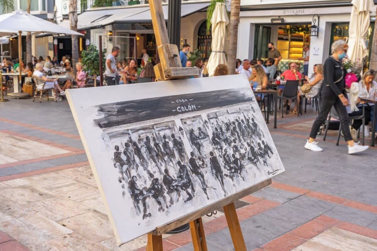 An outdoor café with tables and chairs, a wooden easel with a painting, and people dining and walking around.