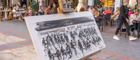 An outdoor café with tables and chairs, a wooden easel with a painting, and people dining and walking around.