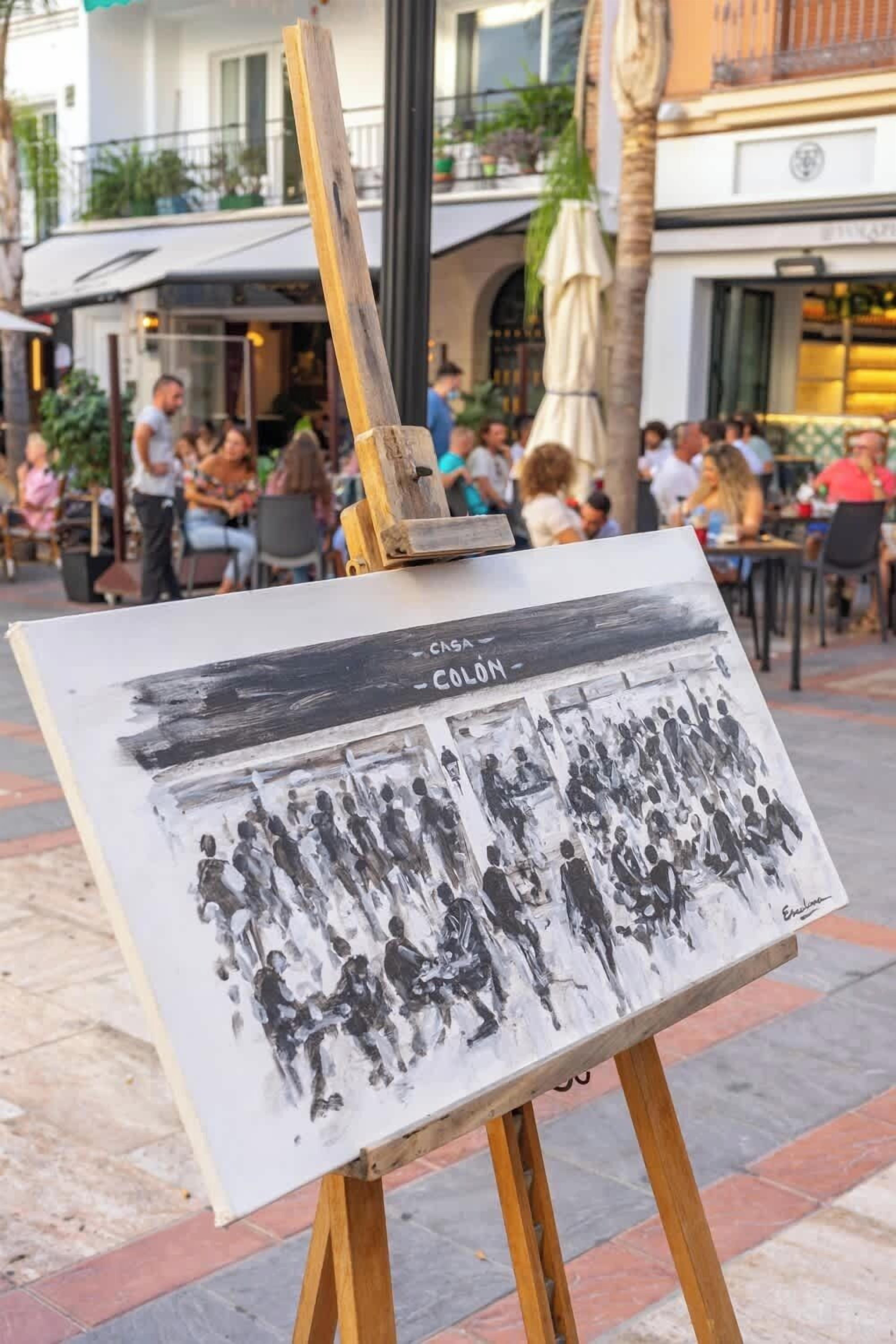 An easel with a black and white painting titled "Colón" in a public outdoor setting.