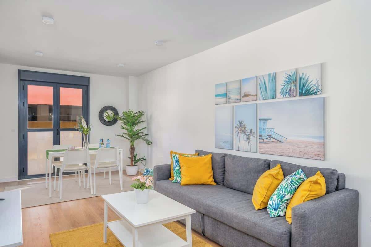 A modern living room with a grey sofa, yellow cushions, a white coffee table, and a dining area with white chairs and a table.