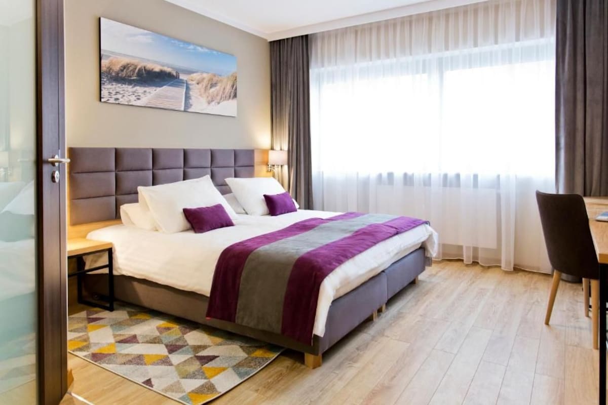A modern hotel room with a large bed, a desk, and a view of the beach through the window.