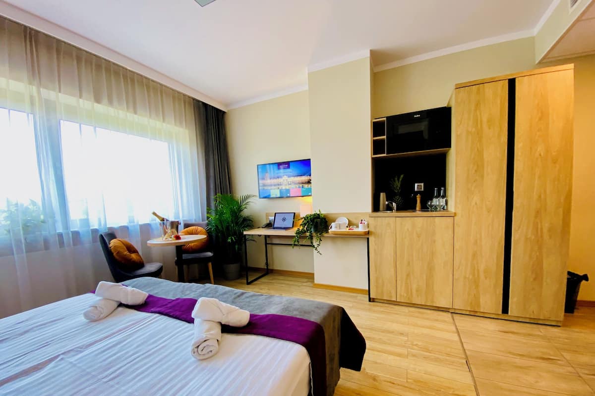 Quarto de hotel com cama, escrivaninha, cadeira, televisão, minibar e uma janela grande.