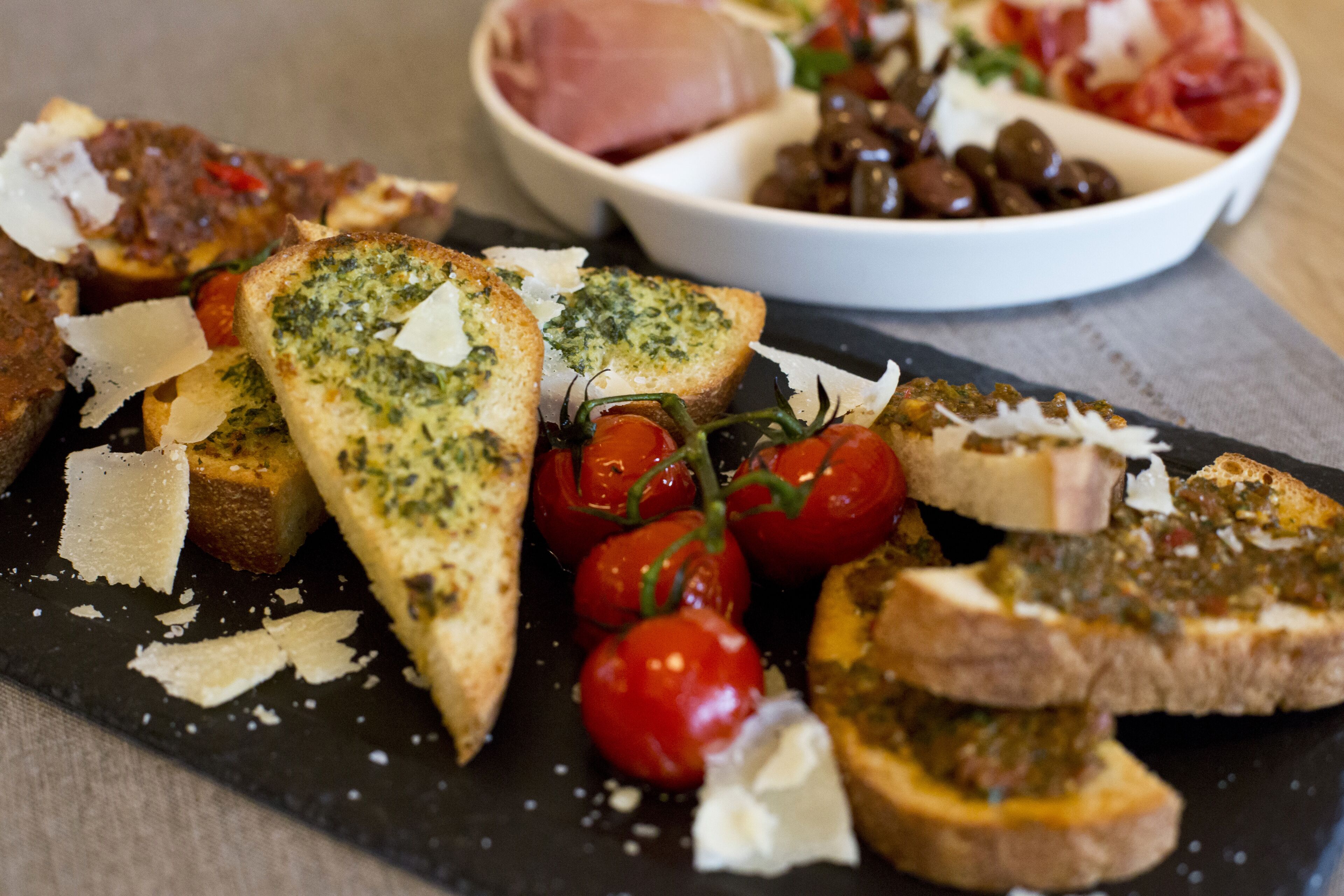 A variety of bruschetta and a bowl of olives and tomatoes on a slate serving board.