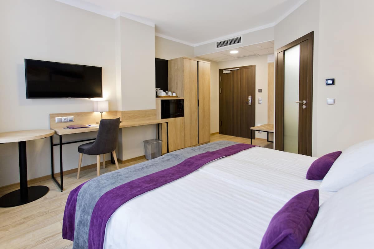 Quarto de hotel com cama, escrivaninha, cadeira, televisão e guarda-roupa.