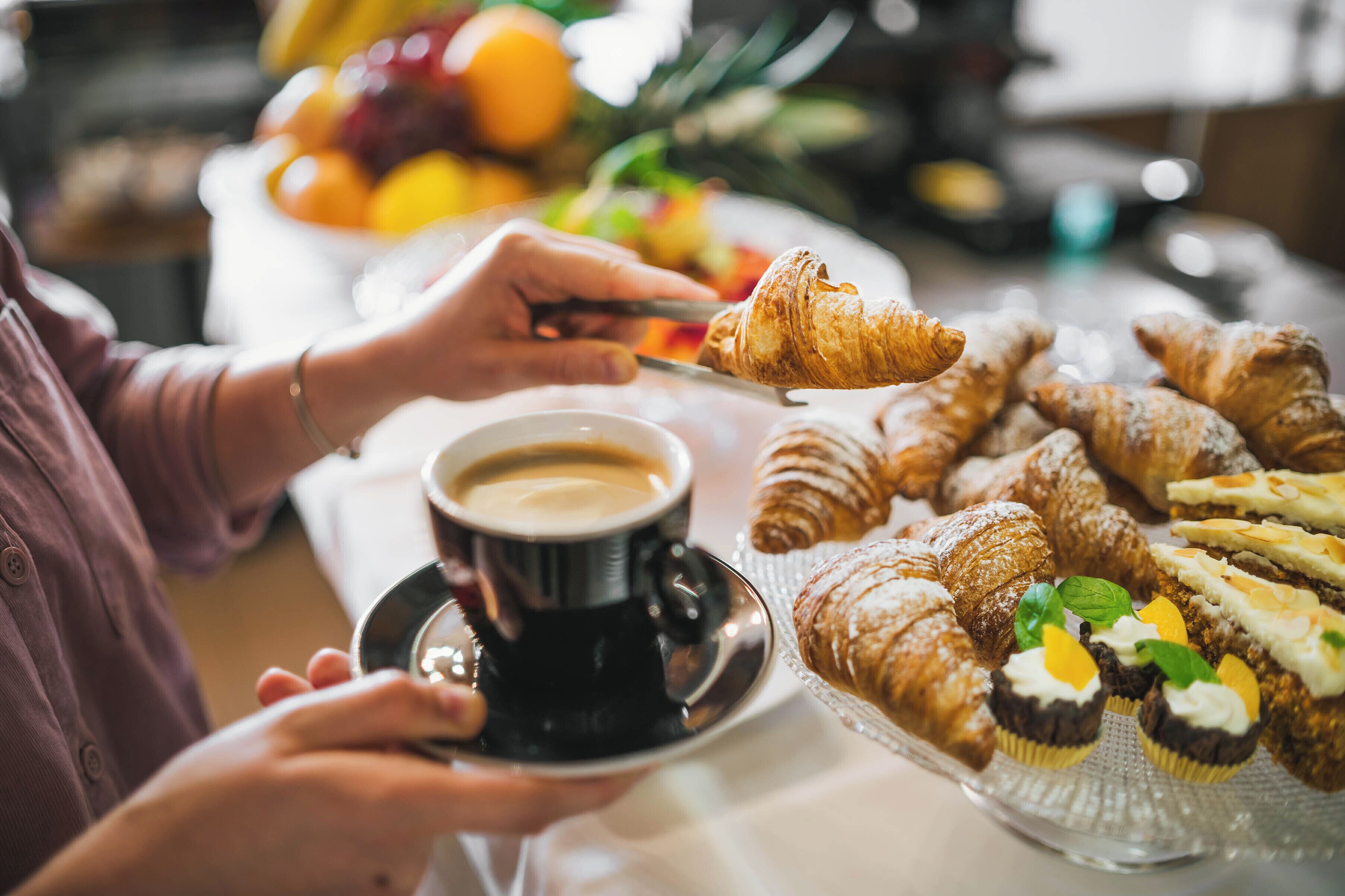 A person holding a croissant and a cup of coffee on a table with various pastries and a bowl of fruit.