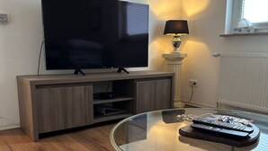 A modern living room with a flat-screen TV on a wooden cabinet, a glass coffee table with a remote control, and a floor lamp. - FeWo Lindenallee – Modern & Ruhig (Nordhorn)
