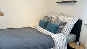 A bedroom with a bed, pillows, a bedside table, and a shelf with a clock. - FeWo Lindenallee – Modern & Ruhig (Nordhorn)