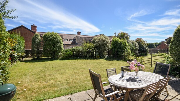 A patio with a round table and chairs, a small garden, and a brick building in the background. - 12 The Gardens (Lyme Regis)