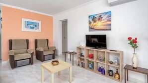 A living room with two armchairs, a TV, a wooden shelf with decorative items, and a painting of a sunset.