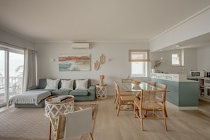 A modern living room with a sofa, dining table, and chairs, a kitchen area with a bar, and a large window.