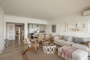 Un salon avec un canapé, des fauteuils, une table basse et, au fond, une cuisine.