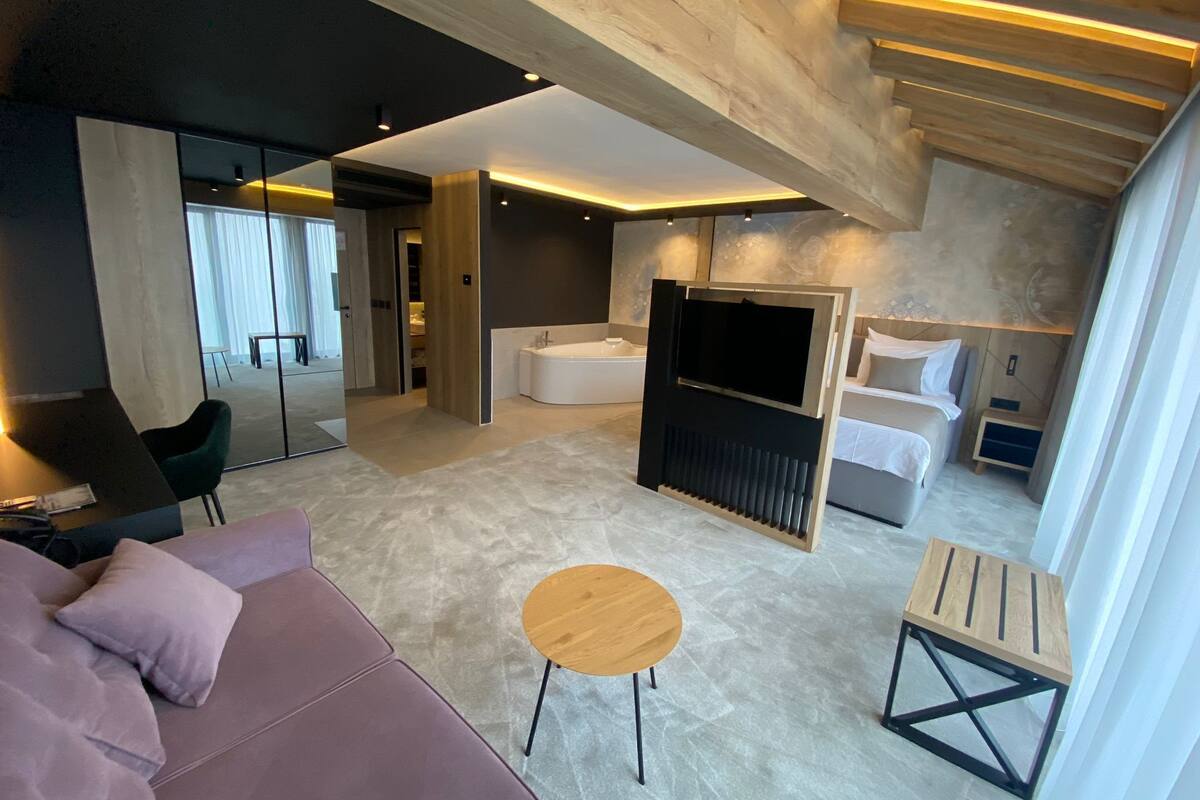 A modern hotel room with a sofa, a small round table, a bed, a TV, and a bathroom.