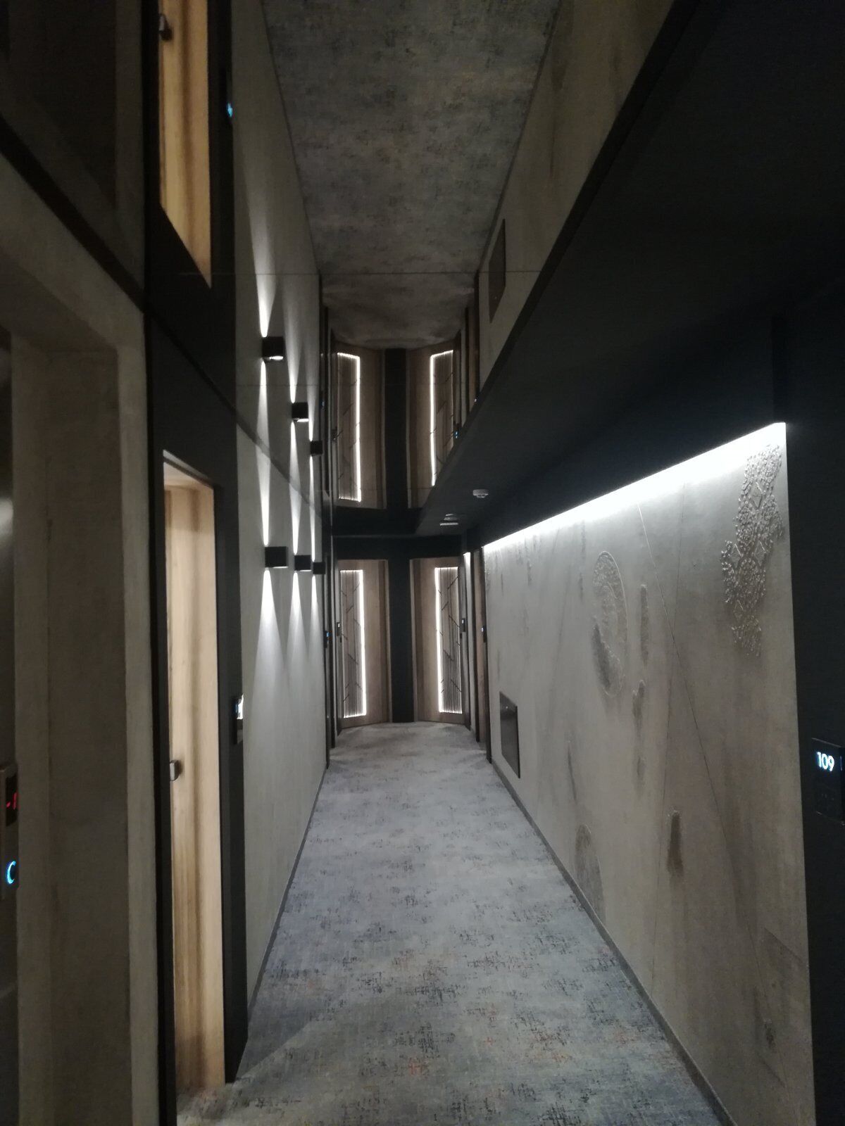 A narrow, dimly lit hallway with a concrete floor and walls, flanked by doors and illuminated by vertical light strips.