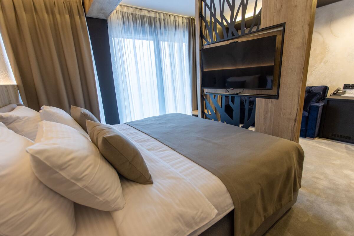 A hotel room with a large bed, a TV mounted on the wall, and a window with curtains.