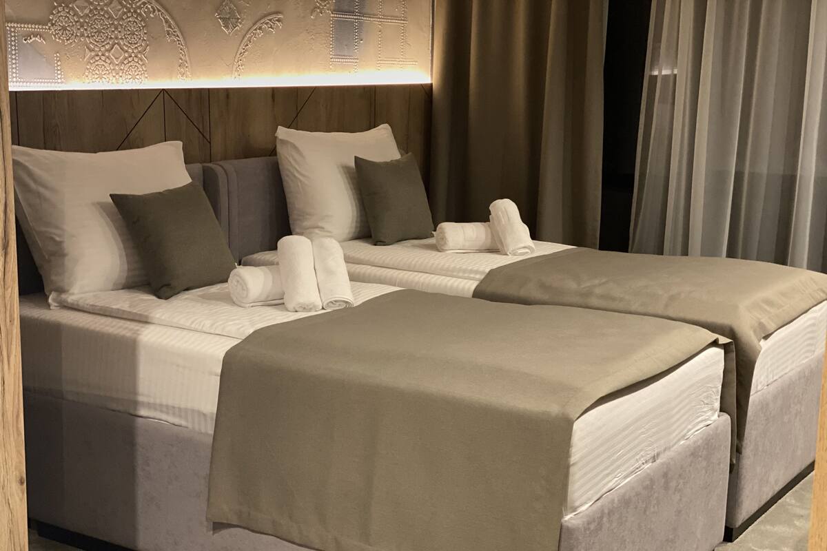 A hotel room with two beds, each with a white comforter and pillows, a wooden headboard, and a wall-mounted light fixture.