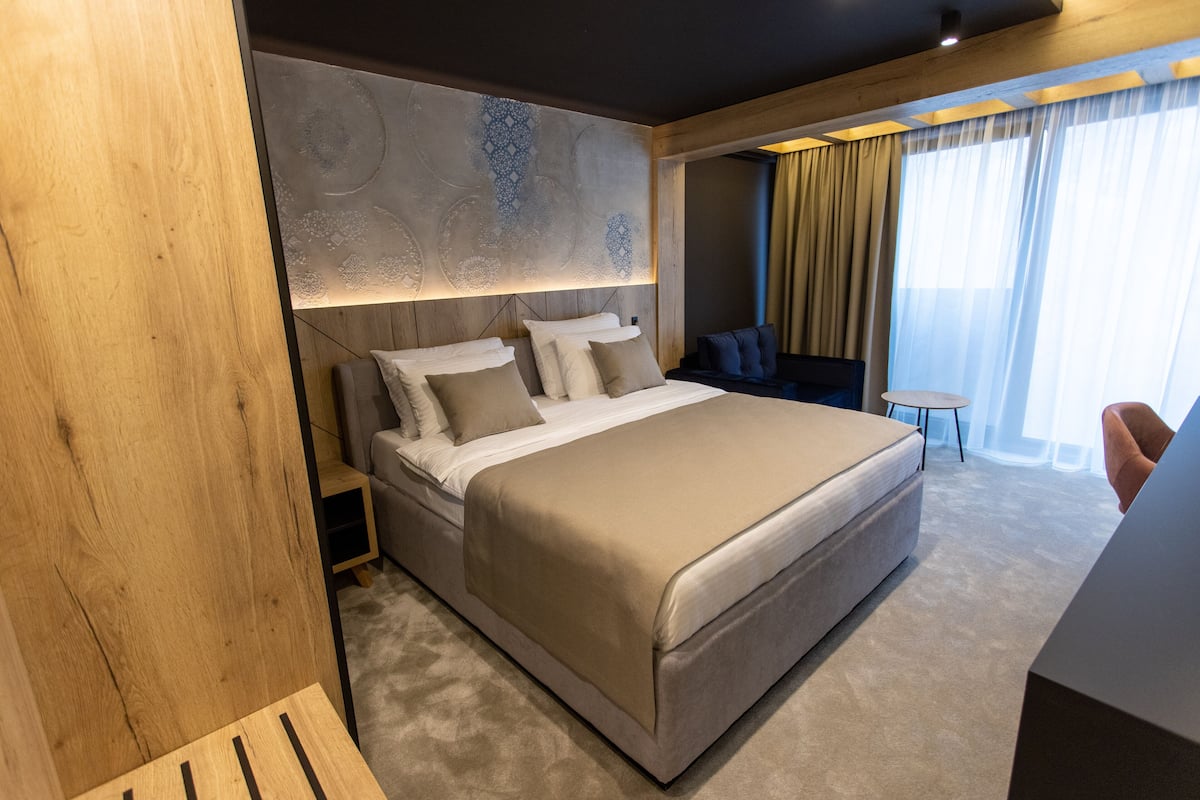 A modern hotel room with a large bed, a wooden accent wall, and a view of the city through large windows.