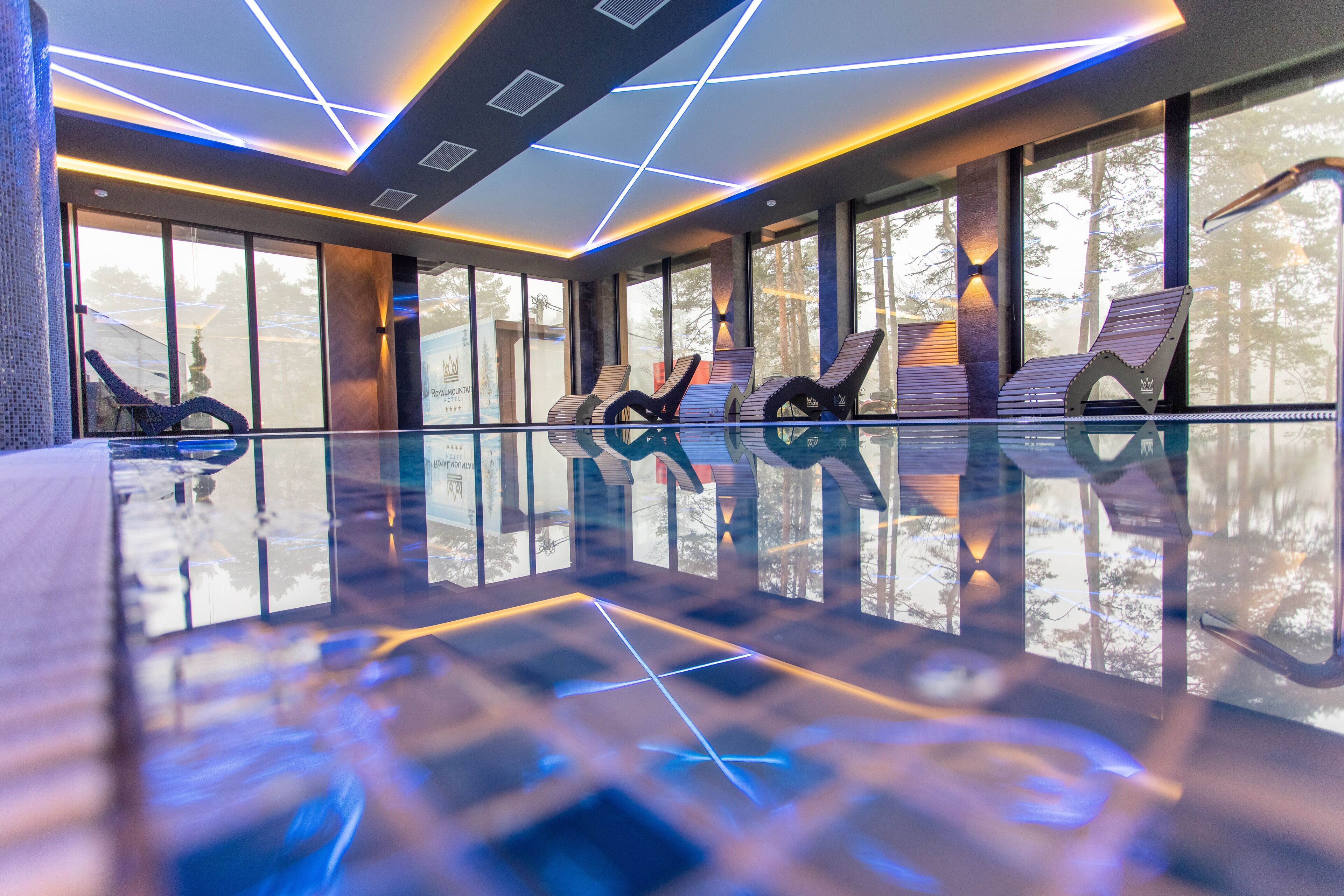 Ein moderner, überdachter Poolbereich mit geometrischer Deckenbeleuchtung, großen Fenstern und außergewöhnlichen Liegestühlen.