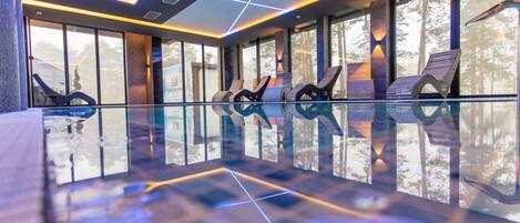 A modern indoor pool area with geometric ceiling lighting, large windows, and unique lounge chairs.