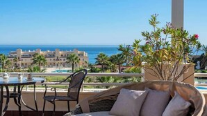 Un sofá de mimbre con cojines en una terraza con vista a una piscina y al océano, con una mesa y sillas cerca.