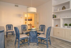 Un espace repas avec une table à plateau en verre, entourée de chaises bleues, un luminaire suspendu blanc et un meuble blanc avec des étagères.