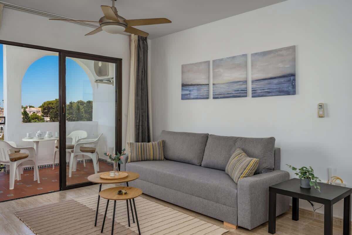 Ein modernes Wohnzimmer mit grauem Sofa, einem kleinen Tisch und einem Deckenventilator. An der Wand hängen drei Gemälde und durch eine Glastür bietet sich ein Blick ins Freie.