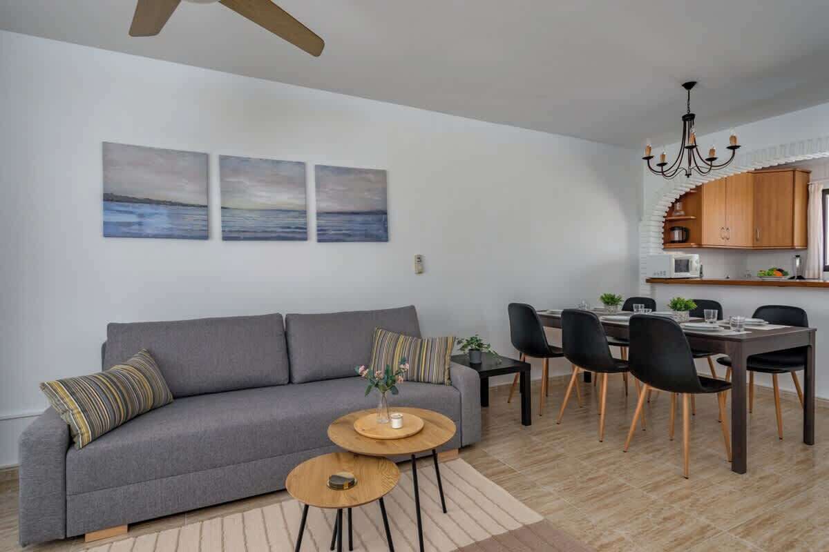 A modern living room with a grey sofa, a dining table with chairs, and a kitchen area with wooden cabinets and a chandelier.