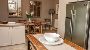 A kitchen with a stainless steel refrigerator, wooden dining table, and chairs. There is a vase with flowers on the table.