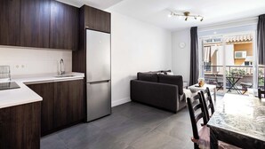 A modern kitchen with dark wood cabinets, a stainless steel refrigerator, and a white countertop. There is a dining area with a glass table and chairs, and a sofa in the background.