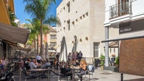 An outdoor café with tables and chairs, a building with a sign reading "EL CIELO", and palm trees in the background.