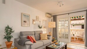 A living room with a grey sofa, a glass coffee table, and a potted plant. There are framed pictures on the wall and a window with curtains. - Beachfront Apartment w/ Pool & Golf Access - La Duquesa (Málaga)