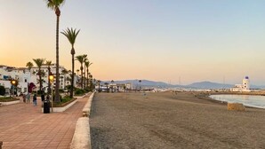 A beach with a paved walkway lined with palm trees and street lamps, leading to the water. - Beachfront Apartment w/ Pool & Golf Access - La Duquesa (Málaga)
